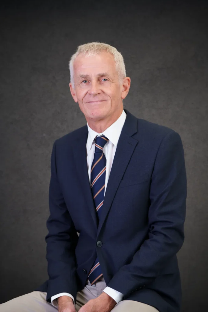 Our Team | Rugby School Nigeria An older man with short grey hair wearing a navy suit, white shirt, and striped tie sits and smiles slightly in front of a dark grey textured background—an image that captures the calm confidence of Benedict Tucker.