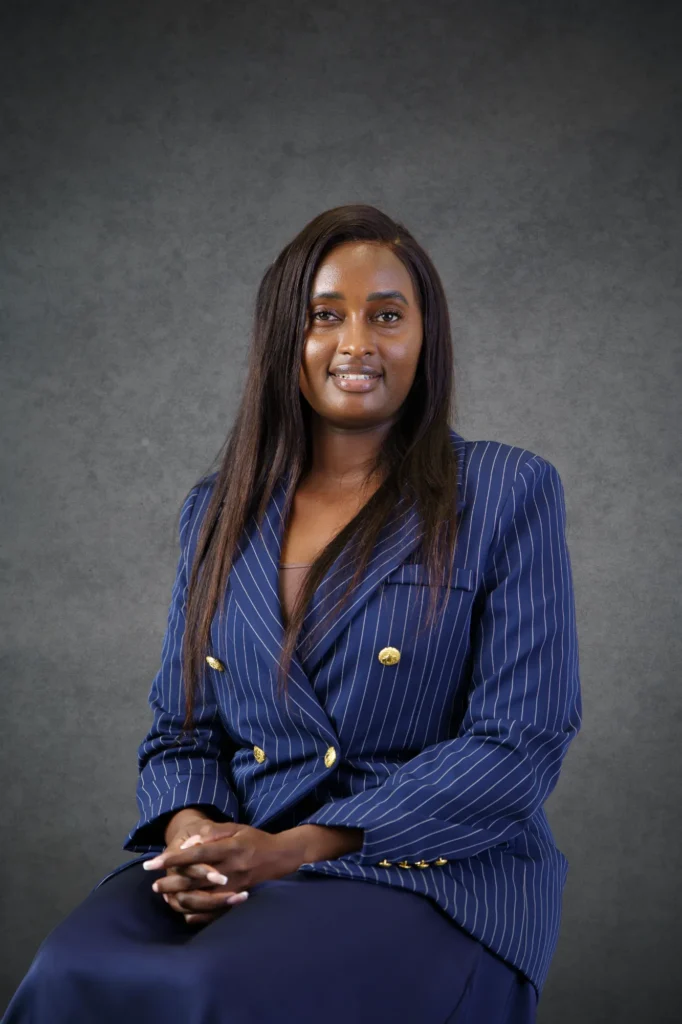 Our Team | Rugby School Nigeria A woman with long hair wearing a blue pinstripe suit sits in front of a grey textured background, hands clasped in her lap, and smiles softly at the camera—Caroline Njilu embodies both confidence and warmth in this poised portrait.
