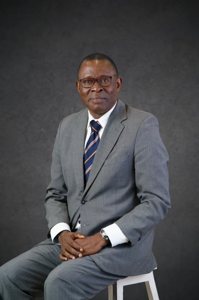 Our Team | Rugby School Nigeria Charles Anumokwu, dressed in a grey suit and striped tie, sits on a stool against a dark grey background, smiling slightly with his hands resting on his knees.