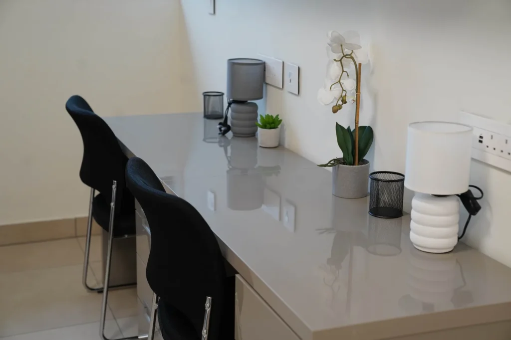 A Home Away from Home | Rugby School Nigeria A modern workspace features two black chairs at a sleek, grey desk with small potted plants, a white lamp, a mesh pen holder, and a decorative orchid against a white wall with power sockets.