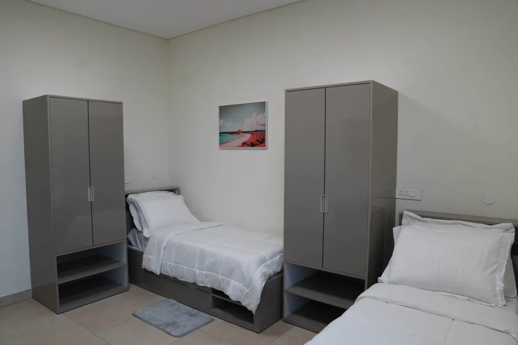 A Home Away from Home | Rugby School Nigeria A clean, minimalist room with two single beds, each with white bedding, separated by two grey wardrobes. A small mat is on the floor, and a beach photograph hangs on the cream coloured wall above one bed.