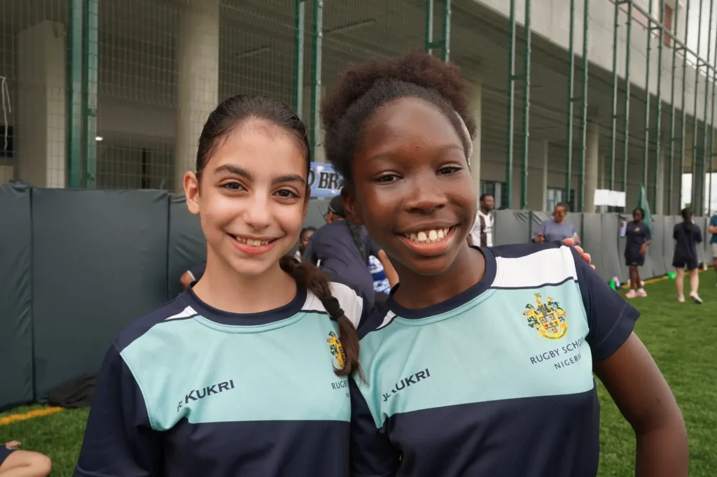 Welcome to Rugby School Nigeria | Rugby School Nigeria Two girls in Rugby School Nigeria jerseys smile at the camera whilst standing on a green sports field, with other pupils and a large building visible in the background.