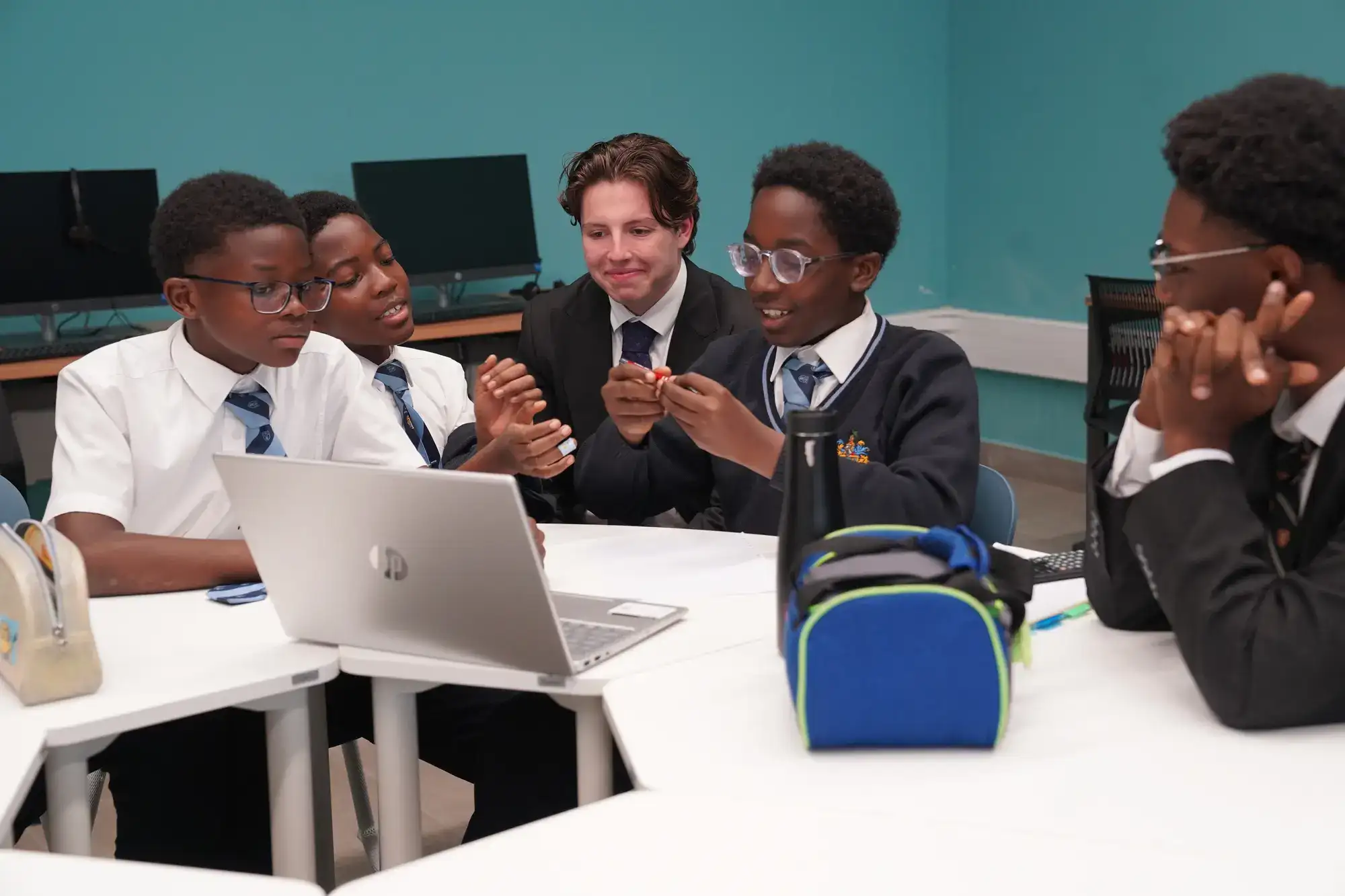 Five students in school uniforms sit around a laptop, engaged in discussion. They appear to be working on a group project in a classroom with computers and blue walls, reflecting the collaborative learning environment at Rugby School Nigeria.