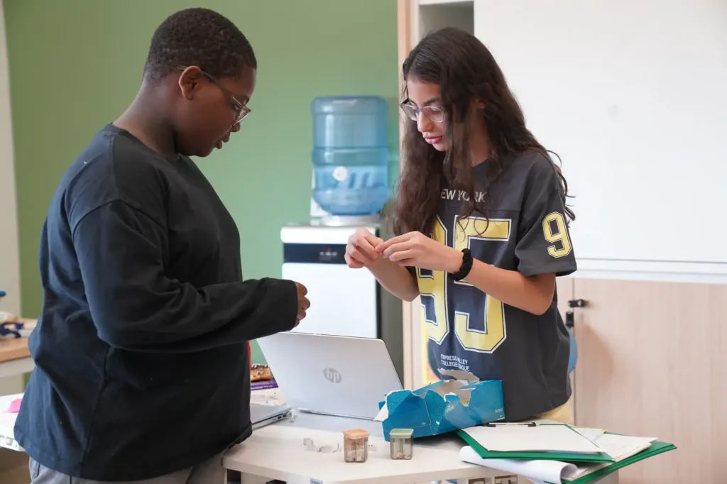 Two students stand beside a desk with a laptop, exploring small containers as part of an Adaptive Learning activity. One holds an object while the other watches attentively. Folders, a water cooler, and a bag are scattered on the desk in this dynamic classroom setting.