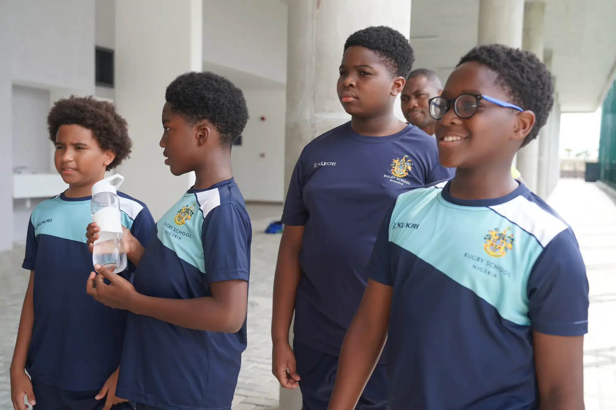 Four boys in blue sports kits stand together outside, one holding a water bottle. Appearing relaxed near large columns with a partly visible building and brick paving in the background, these Key Stage 3 pupils share a moment between activities.