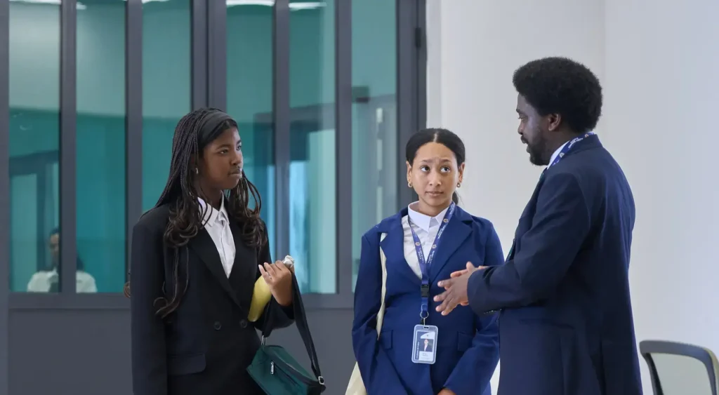 Three people in business attire stand indoors, having a conversation. Two women listen whilst a man gestures as he speaks. All wear ID badges.