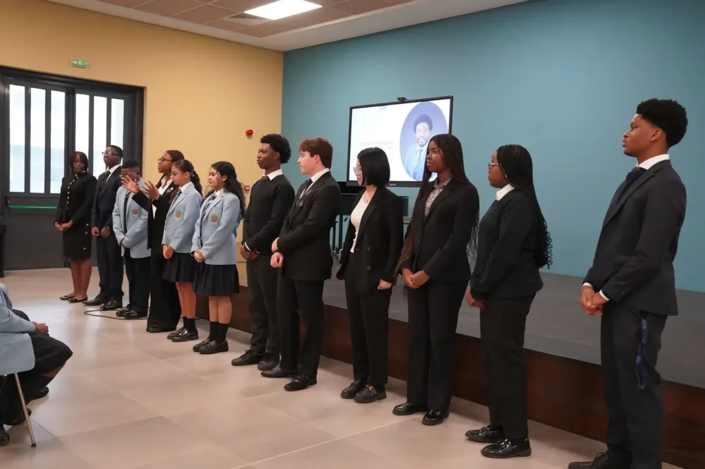 Key Stage 5 (Sixth Form) | Rugby School Nigeria Twelve Sixth Form students in formal attire stand in a line facing an audience in a school hall, with a screen displaying a photo and text behind them.