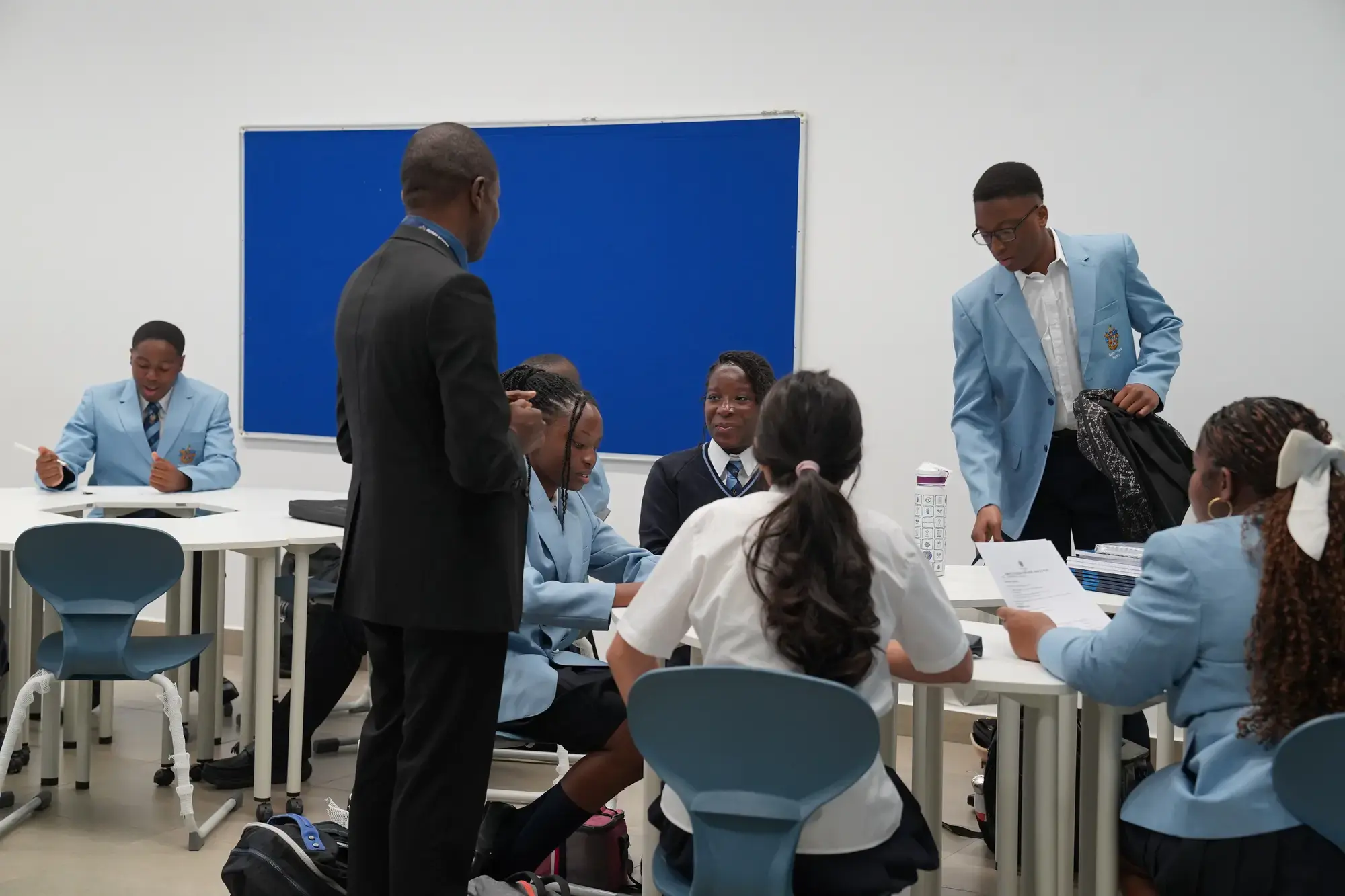 A teacher stands by a group of students in blue uniforms sitting at desks, whilst another student in a blazer stands holding papers. The classroom, designed to support adaptive learning, features white walls and a blank blue noticeboard.
