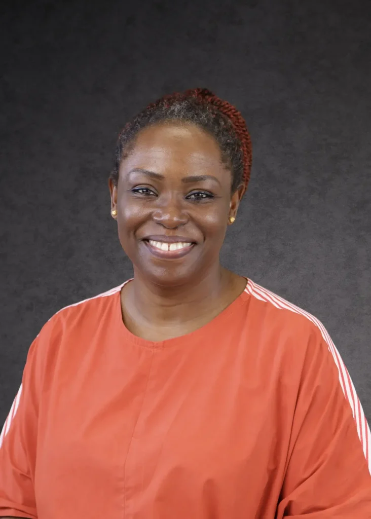 Our Team | Rugby School Nigeria A woman with short, curly hair pulled back, wearing an orange top with white stripes on the sleeves, smiles in front of a dark grey textured background.