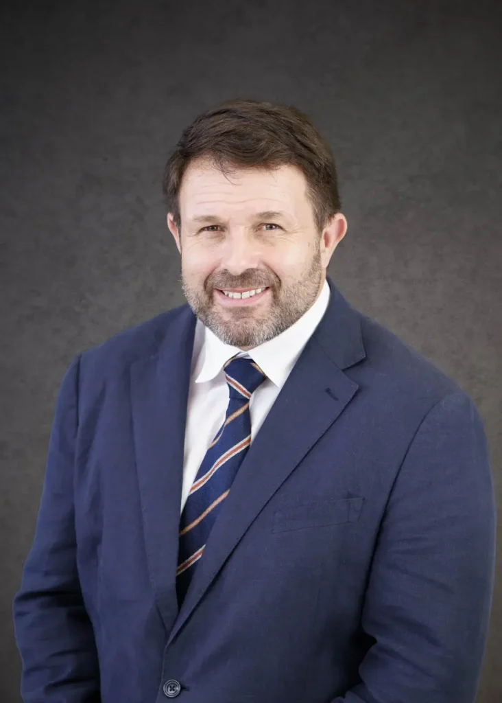 Our Team | Rugby School Nigeria A man with short brown hair and a beard wears a navy suit, white shirt, and striped tie, standing against a dark grey background.