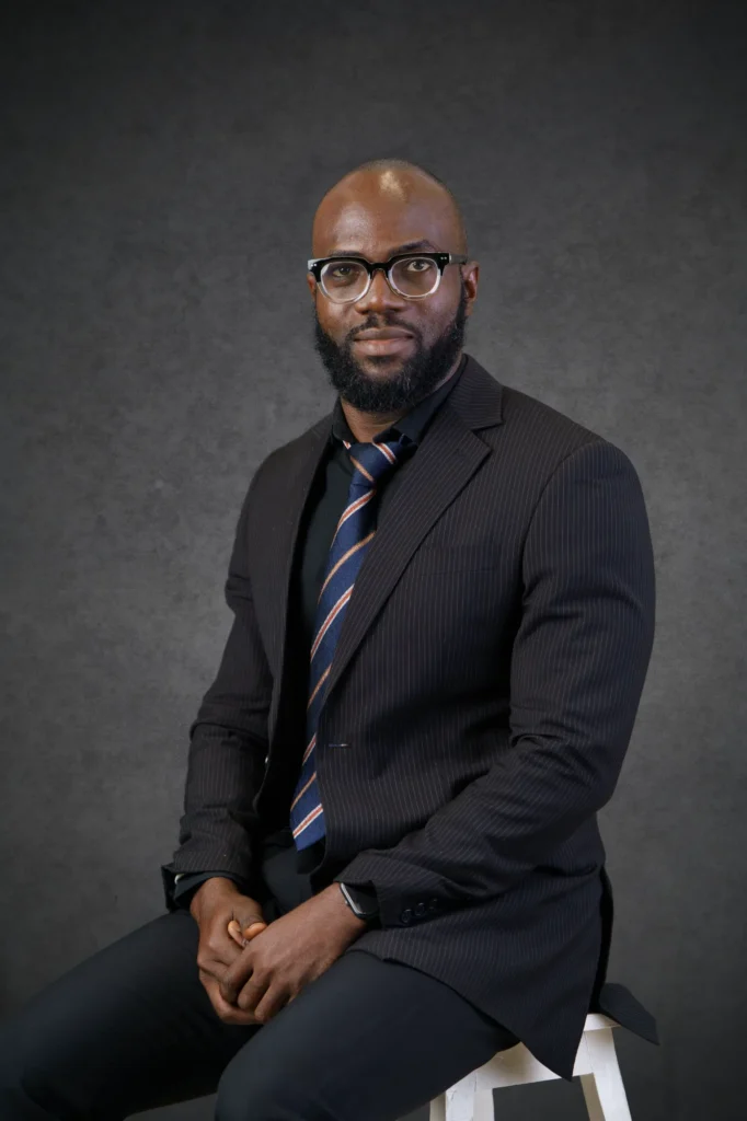 Our Team | Rugby School Nigeria Elijah Odewunmi, dressed in a dark pinstripe suit, blue striped tie, and glasses, sits on a white stool against a grey textured background, looking confidently at the camera.