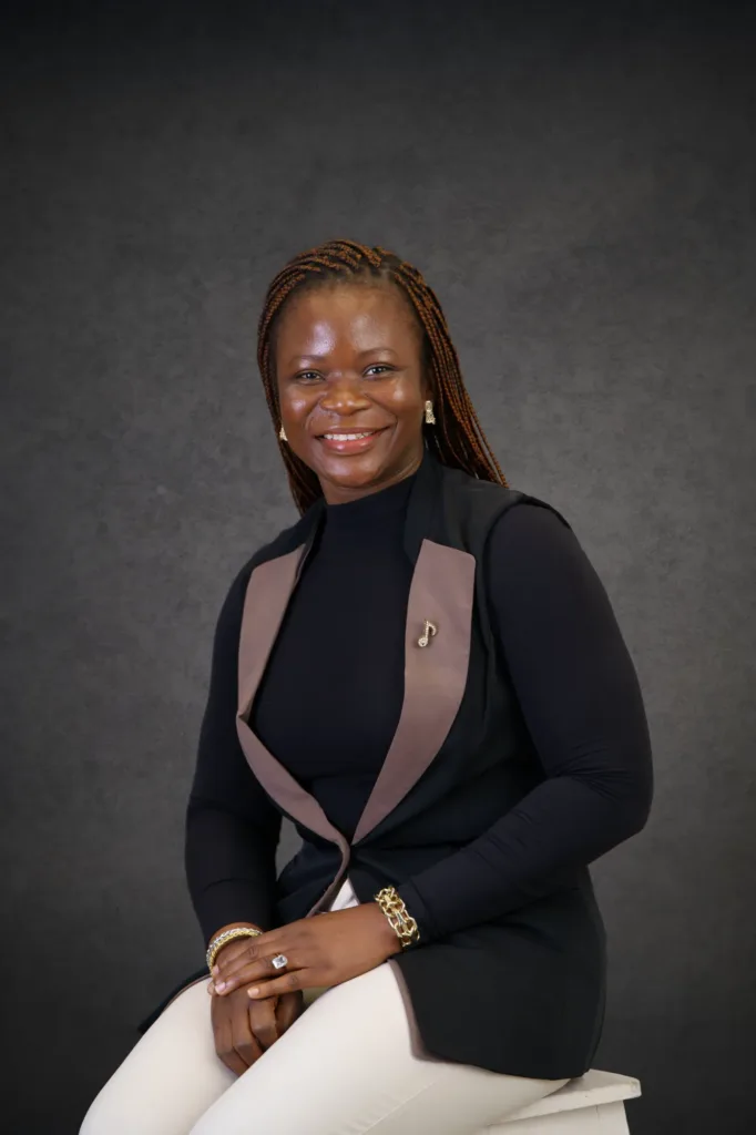 Our Team | Rugby School Nigeria Sandra Onoja, a woman with braided hair, wearing a black long-sleeve top, black waistcoat with taupe lapels, and light trousers, smiles whilst seated on a stool against a dark grey studio background.