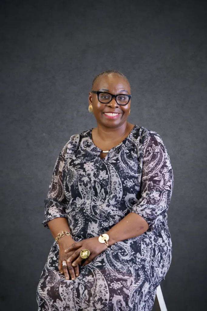 Our Team | Rugby School Nigeria Smiling woman with short hair and glasses, wearing a patterned dress and gold jewellery, sits on a stool in front of a dark, textured background—Danielle McGuinness exudes confidence and warmth.
