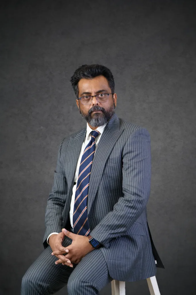 Our Team | Rugby School Nigeria A man with glasses and a beard, wearing a grey pinstripe suit, white shirt, and striped tie, sits on a stool against a dark grey studio background.