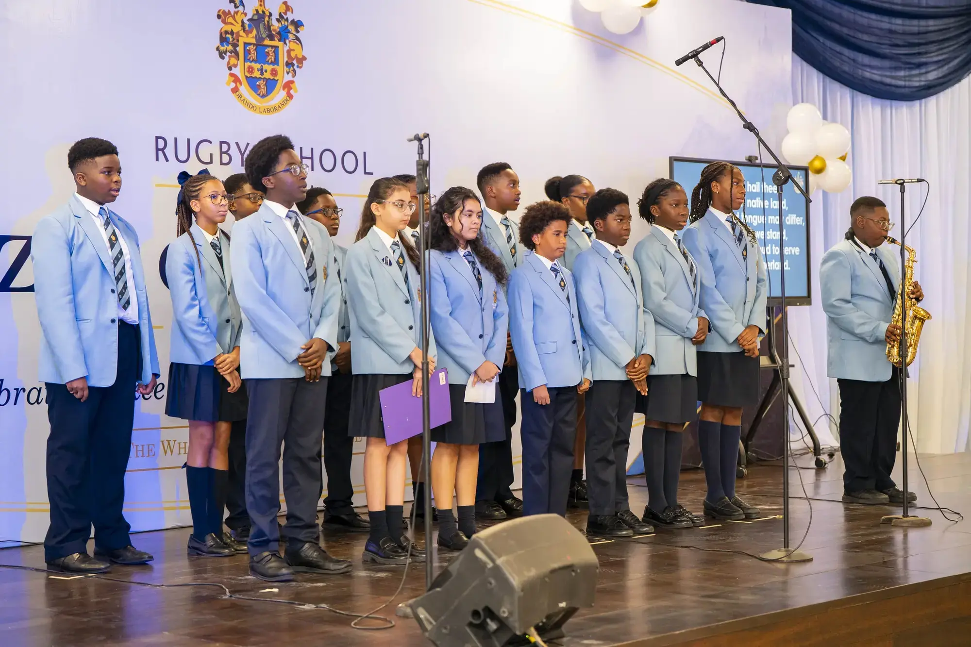 A group of Sixth Form students in blue blazers and ties stand on stage at Rugby School, some singing, one holding a saxophone, with microphones and a projection screen in the background.