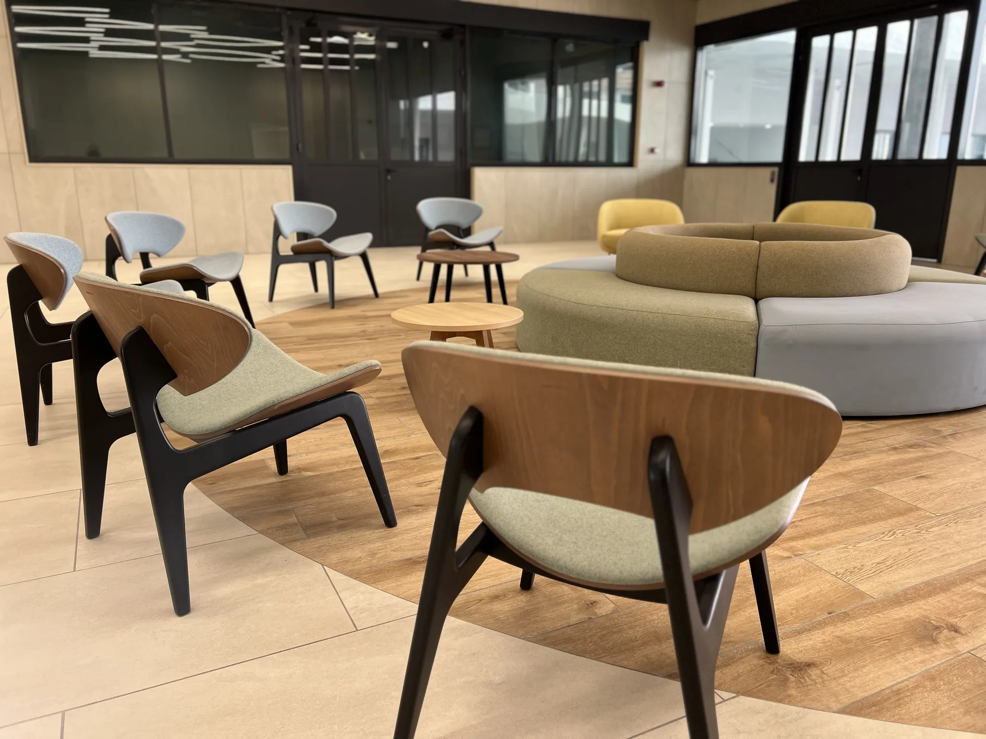 A modern room with wooden and upholstered chairs arranged in a circle, featuring a round, cushioned seating area in the centre on a floor with wood and beige tile patterns. Large windows and glass doors line the walls.