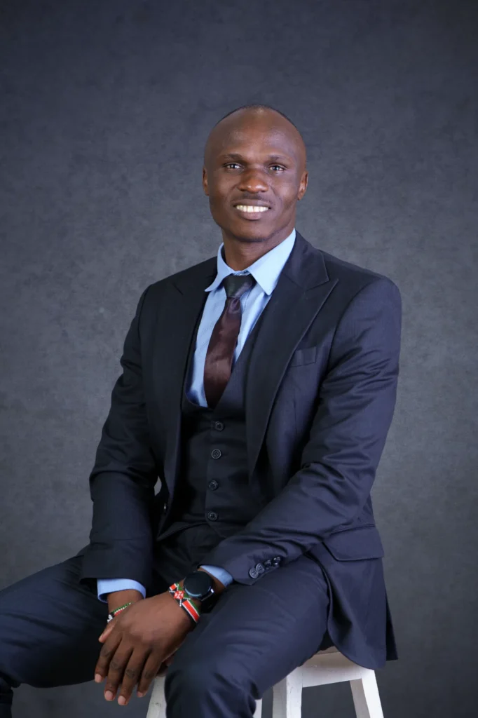 Our Team | Rugby School Nigeria A man in a dark suit, blue shirt, and tie sits on a stool against a grey background, smiling at the camera. Kennedy Ombuni wears a wristwatch and a beaded bracelet.