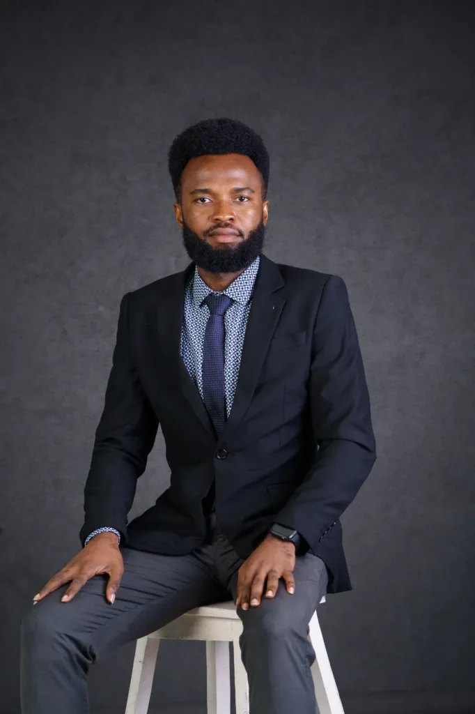 Our Team | Rugby School Nigeria A man, Omotayo Oluwaseyi, in a black suit, blue patterned shirt, and tie sits on a stool against a dark grey background, looking at the camera with a neutral expression.