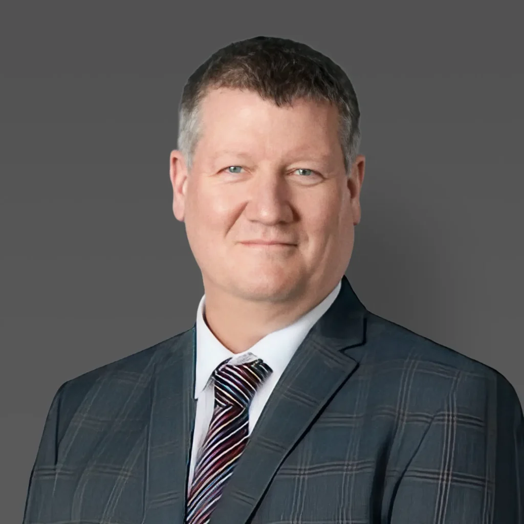 Our Team | Rugby School Nigeria A middle aged man with short brown hair, wearing a dark checked suit, white shirt, and striped tie, poses in front of a plain grey background, smiling slightly—introducing Gareth Nelson.